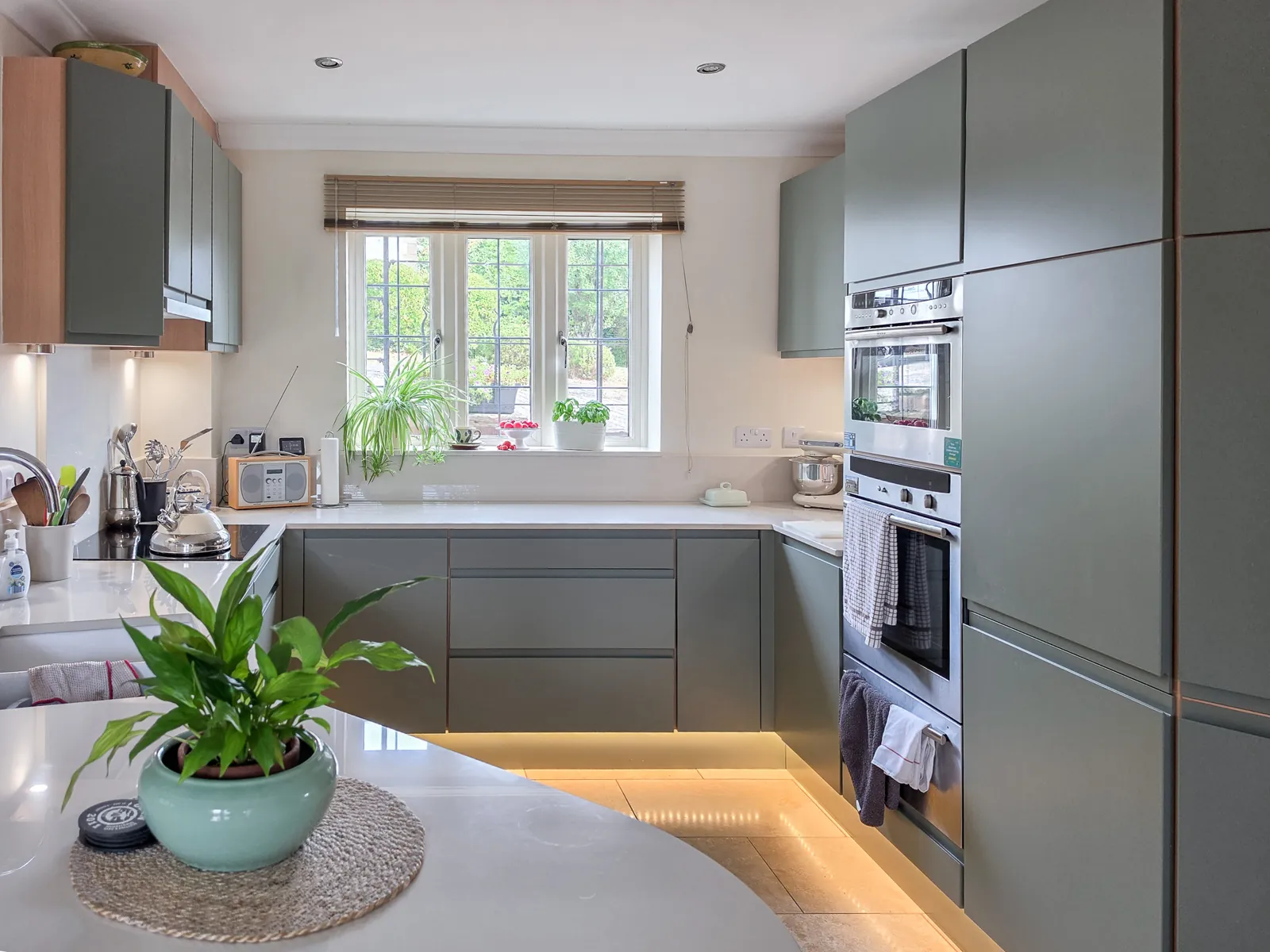 The finished Brinkworth kitchen with handleless green units, a curved worktop in the foreground and a bright window